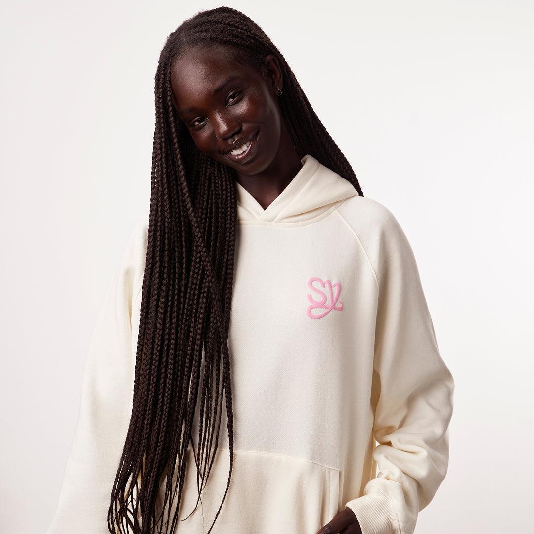 XS | S | M | L | XL | XXL |Woman wearing a cream-colored hoodie with a pink logo on a white background