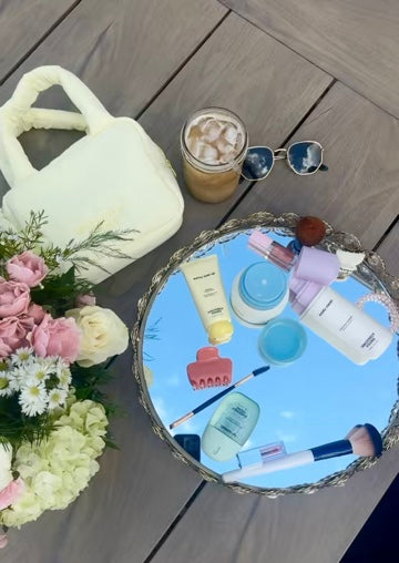 Cosmetic items on a round mirror with a cup, sunglasses, and flowers on a wooden surface