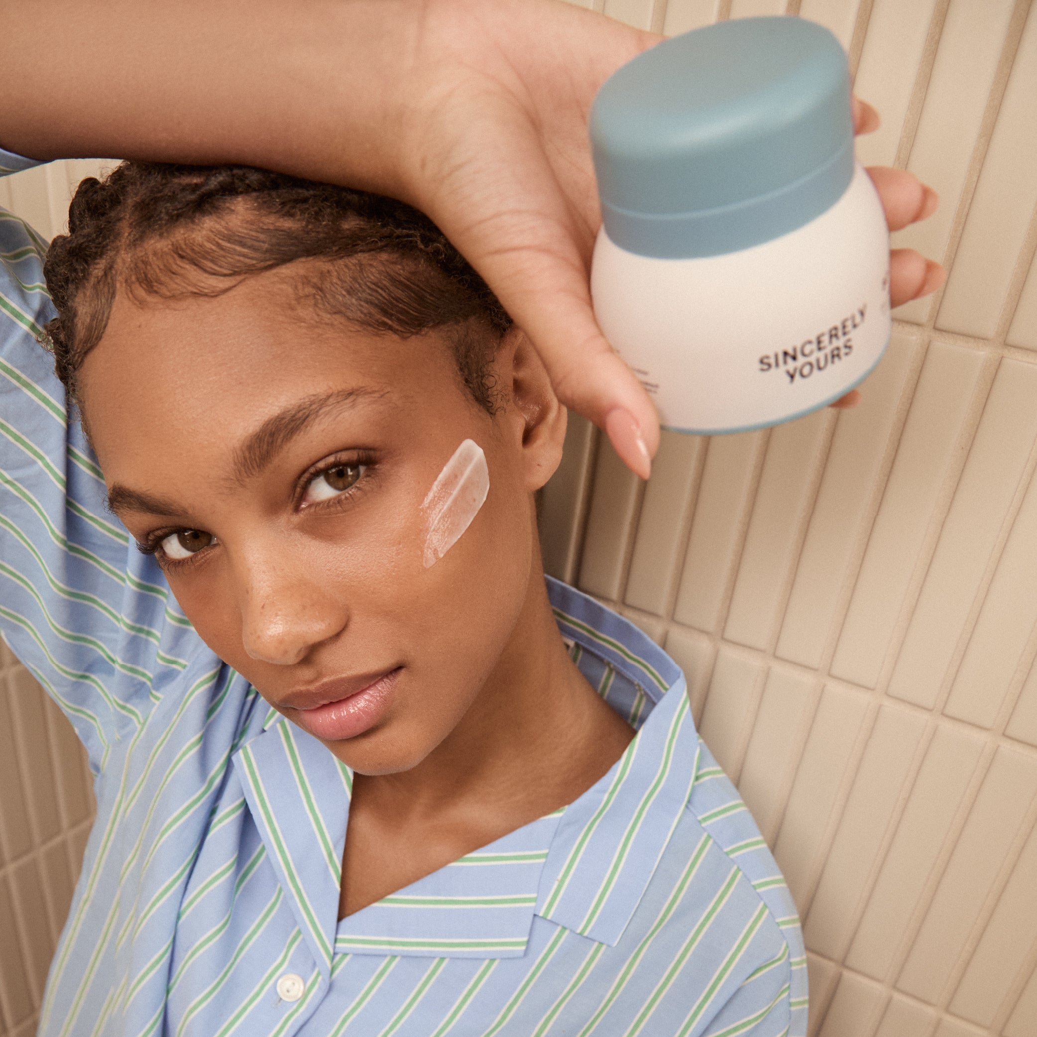 Person applying cream to face with 'Sincerely Yours' jar in hand, tiled bathroom background